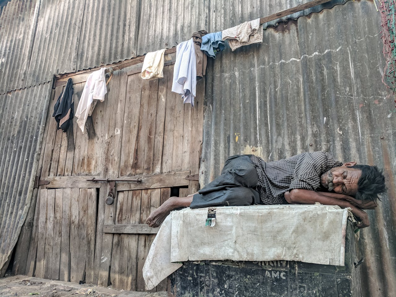 An elderly man peacefully sleeps against a rundown wooden wall with hanging clothes.