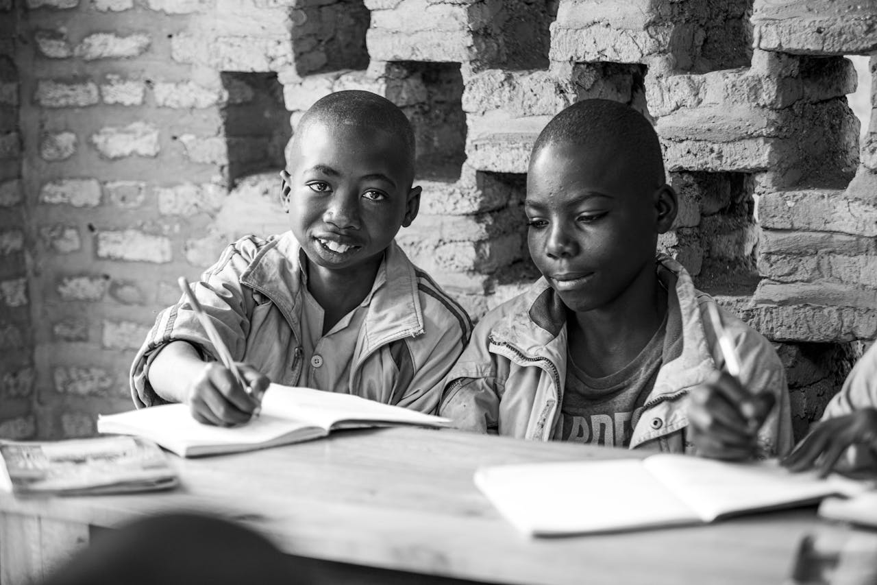 Black and white photo of two children studying with notebooks and pens against a brick wall.