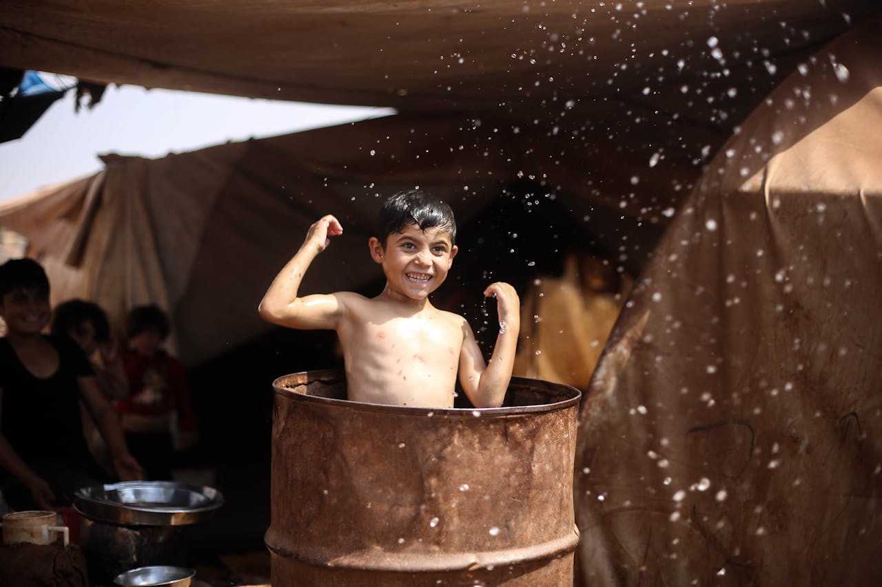A smiling boy splashes water in a barrel under tents, showcasing joyful moments in a refugee camp.